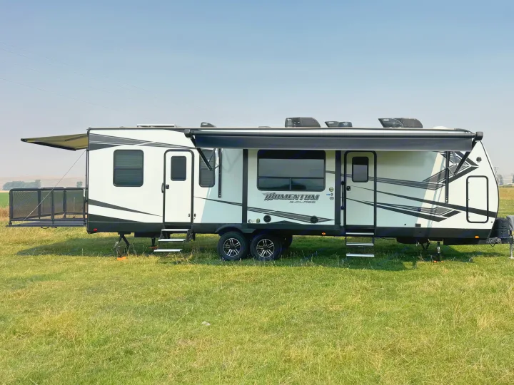 A white Momentum G-Class toy hauler travel trailer rv rental is set up in a grassy field with awnings extended.. Grand Design Momentum 2022