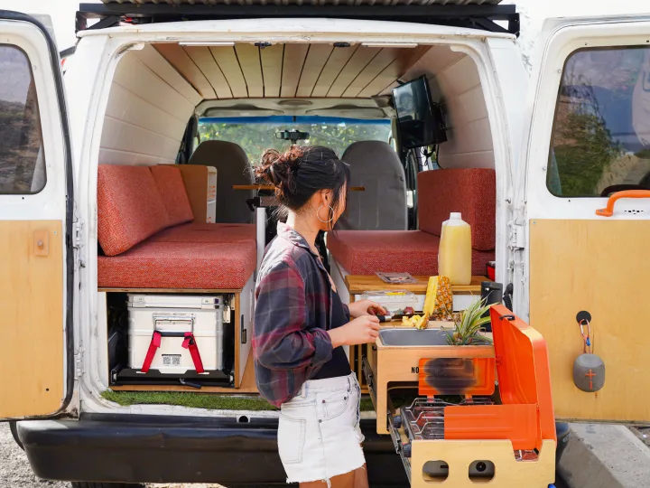 RV rental camper van with woman preparing pineapple in its slide-out kitchen. Custom interior features a bed, TV, & storage.. Ford Econolline 250s 2005