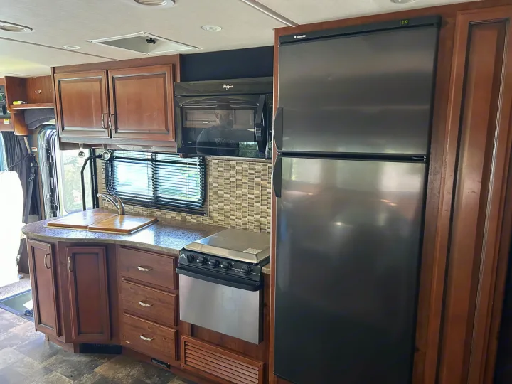 Modern RV rental kitchen featuring dark wood cabinets, stainless steel fridge, microwave, stove, and sink with tiled backsplash.. Bounder Classic 2014