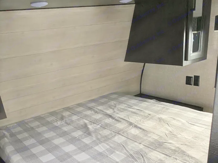 RV rental bedroom with a bed featuring gray plaid sheets, light wooden wall, and dark overhead cabinet.. Jayco Jay Flight SLX 2022