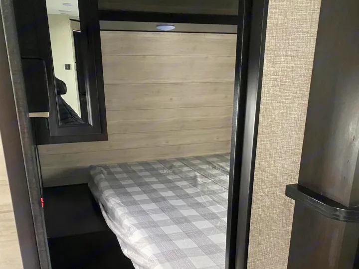 RV rental bedroom: a compact sleeping area featuring a plaid bed, light wood wall paneling, and a framed mirror.. Jayco Jay Flight SLX 2022