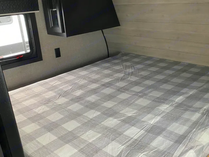 This RV rental bedroom features a cozy bed with a grey plaid cover, a window, and a storage cabinet, ideal for travel.. Jayco Jay Flight SLX 2022