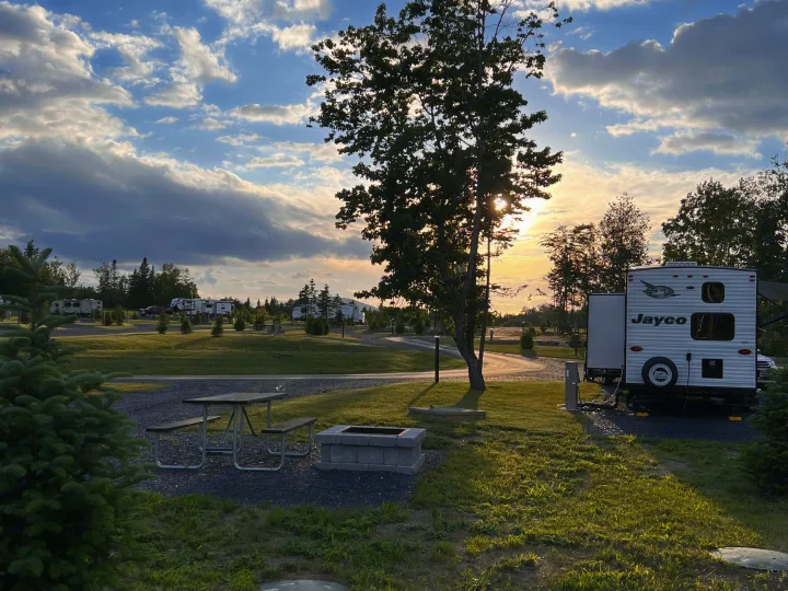 Experience tranquil RV rental camping at sunset, featuring a Jayco RV, picnic area, and fire pit amidst lush trees and cloudy skies.. Jayco Jay Flight SLX 2022