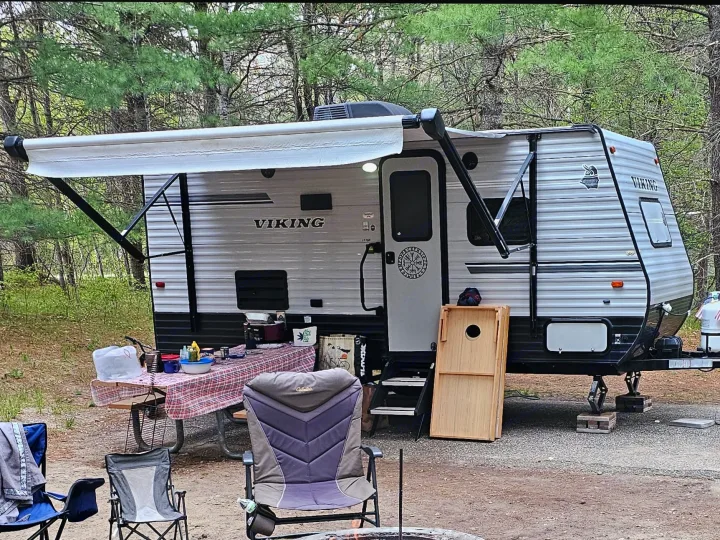 A Viking RV rental at a wooded campsite with an extended awning, picnic table, and chairs set up for outdoor enjoyment.. Forest River Viking 2018
