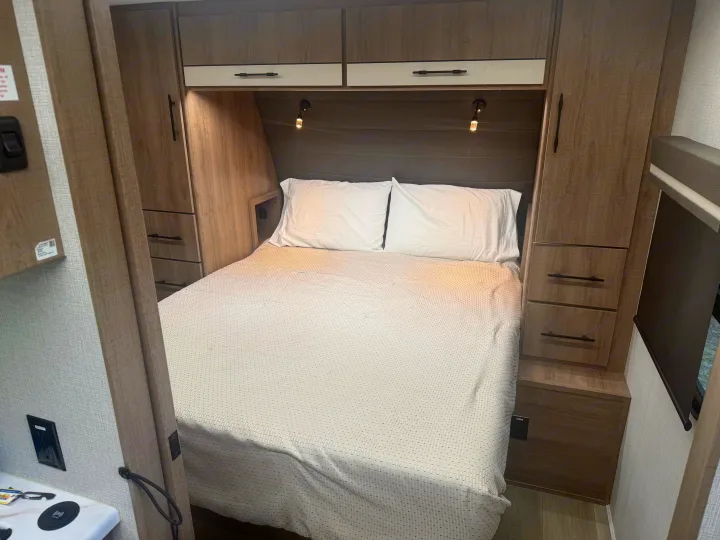 Cozy RV rental bedroom features a comfortable bed, overhead cabinets, and handy side storage with reading lights.. Grand Design Imagine XLS 2024
