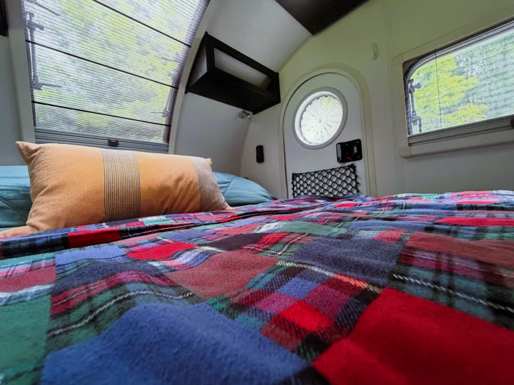 Cozy rv rental bedroom interior, showcasing a bed with a vibrant plaid blanket, accent pillow, and large windows with outdoor views.. nuCamp T@G 2022