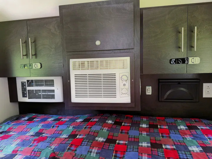 Compact RV rental bedroom featuring dark wood cabinets, two AC units, and a vibrant plaid blanket over the bed.. nuCamp T@G 2022