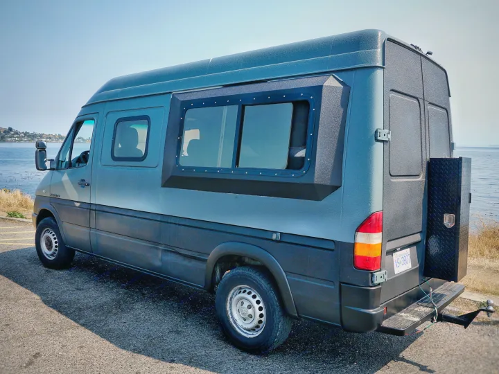 This unique gray camper van, ideal for an RV rental road trip, is parked by the sea with custom windows and a rear toolbox.. Dodge Sprinter 2006