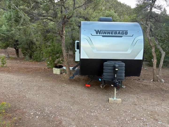 Experience nature with this Winnebago travel trailer RV rental, parked on a gravel campsite amidst tall trees.. Winnebago Access 2025