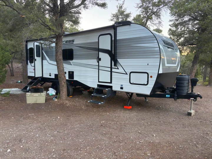This RV rental, a white travel trailer, is parked at a wooded campsite with a cooler and grill setup.. Winnebago Access 2025