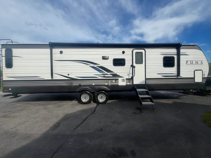 White Puma RV rental travel trailer parked on asphalt under a blue sky. Features extended steps and a rolled-up awning.. Forest River Palomino Puma 2021