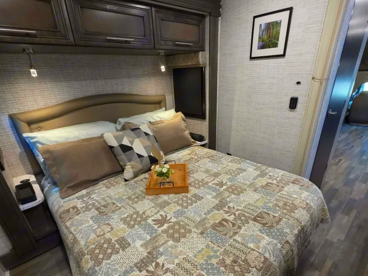 Modern RV rental bedroom with a bed, patterned quilt, overhead cabinets, reading lights, and wall art.. Jayco Greyhawk 2024