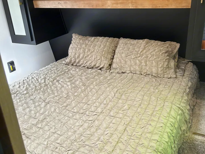 RV rental bedroom with a comfortable bed, textured grey bedding, black headboard, and wood shelf. Compact interior design.. Keystone RV Springdale 2022