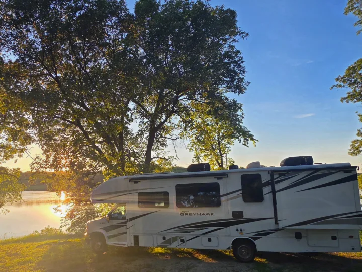 Golden hour camping in an RV rental! A Jayco Greyhawk motorhome is parked beside a serene lake at sunset, surrounded by trees.. Jayco Greyhawk 2024