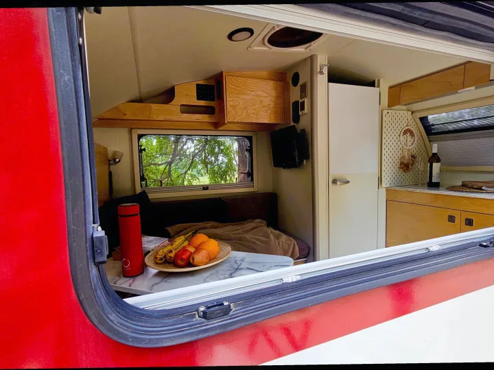 Inside a compact RV rental: a fruit bowl, bed, and small kitchen with a window view of trees.. nuCamp T@B 2022