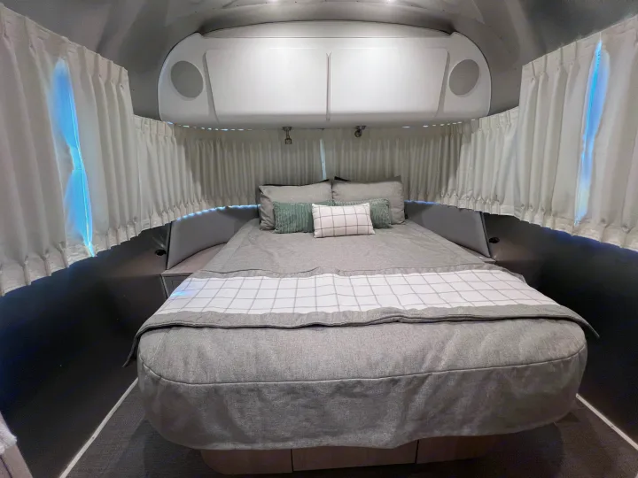 Cozy RV rental bedroom with a comfy bed, modern grey decor, white curtains, and overhead storage, perfect for travel.. Airstream Globetrotter 2022