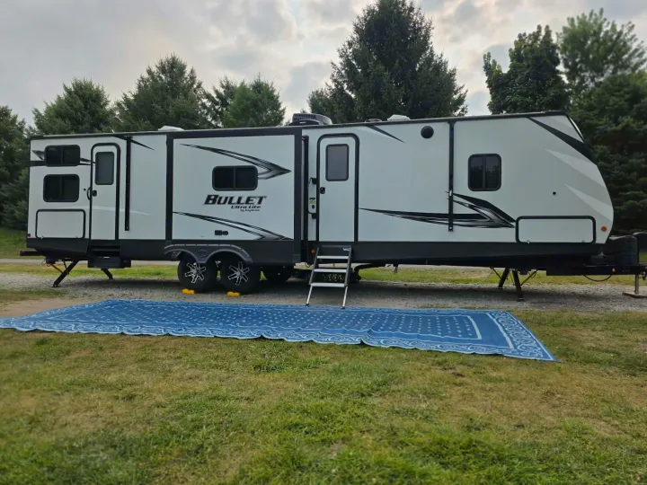 This white Bullet Ultra Lite travel trailer RV rental is set up with a blue mat on a grassy camping site, surrounded by trees.. Keystone RV Bullet Ultra Lite 2021