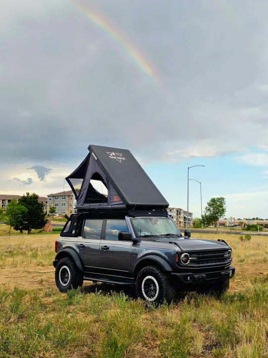 A Ford Bronco with a rooftop tent, parked in a grassy field under a rainbow. Your adventure starts here, ideal for an rv rental.. Ford bronco 2021