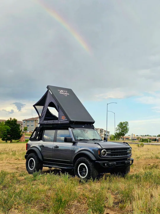 This dark gray Ford Bronco, equipped with a rooftop tent, is perfect for an RV rental outdoor adventure under a vibrant rainbow.. Ford bronco 2021