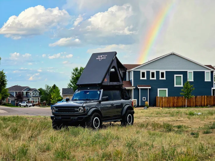 A dark gray Ford Bronco with an open rooftop tent in a grassy field, under a rainbow, clouds & houses. Great for an RV rental adventure.. Ford bronco 2021