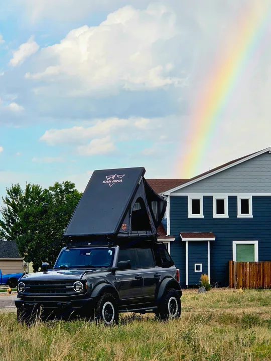 Dark gray Ford Bronco with open SAN HIMA rooftop tent parked by a blue house, a rainbow brightens the sky. Great rv rental for adventure!. Ford bronco 2021