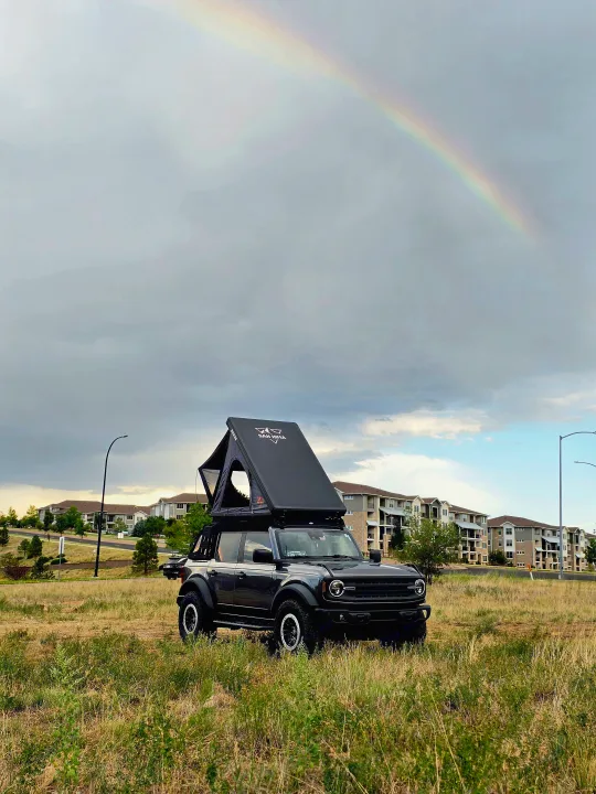RV rental ready: A dark grey Ford Bronco with a rooftop tent in a grassy field under a cloudy sky & rainbow, near buildings.. Ford bronco 2021