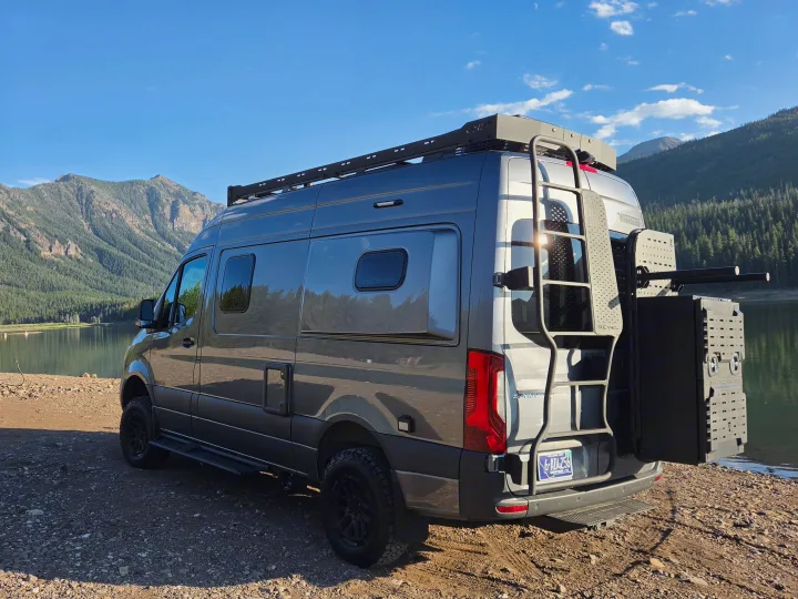 Gray Mercedes Sprinter RV rental camper van with roof rack & ladder, parked by a scenic mountain lake for outdoor adventure.. Winnebago Revel 2025