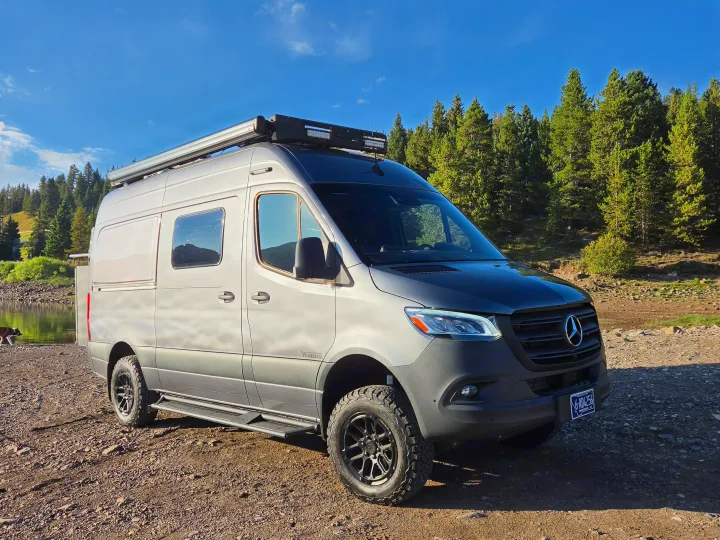 Adventure-ready Mercedes Sprinter camper van for rv rental, parked lakeside with a forest view. A dog walks by the water.. Winnebago Revel 2025