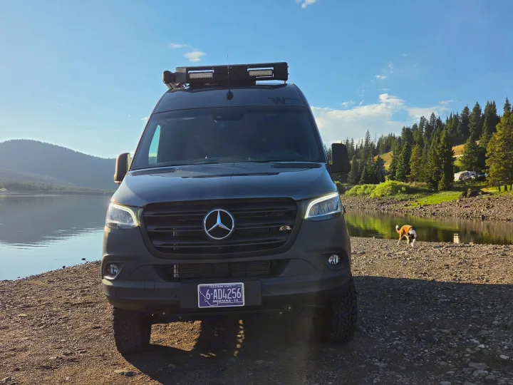 An rv rental Mercedes Sprinter campervan parked on a rocky lake shore with mountains and a dog. Ideal for outdoor adventure.. Winnebago Revel 2025