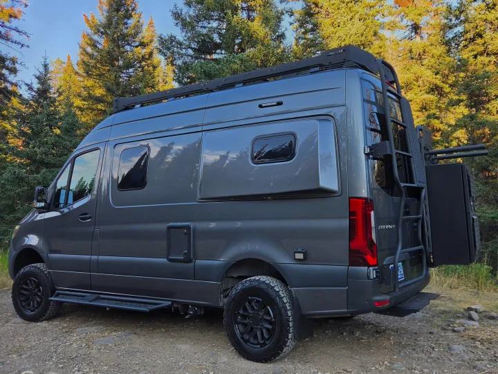 This gray, custom-built camper van, ideal for rv rental adventures, is parked on a dirt path surrounded by evergreen trees.. Winnebago Revel 2025