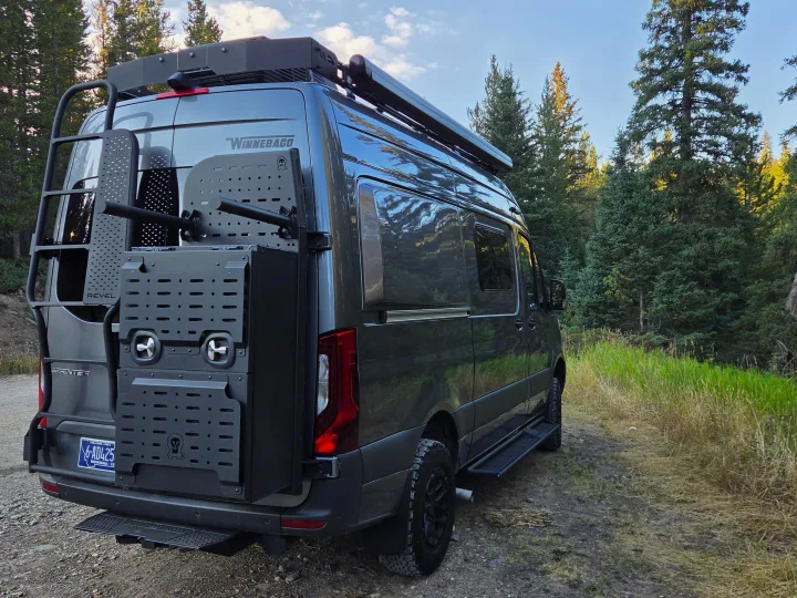 Dark gray Winnebago Revel adventure van, ideal for an rv rental, parked on a dirt road in a forest with a ladder & gear mounts.. Winnebago Revel 2025
