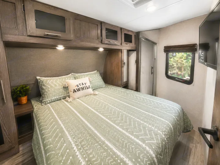 This cozy RV rental bedroom features a comfy bed with green bedding, overhead cabinets, and a window view.. Forest River Surveyor 2018