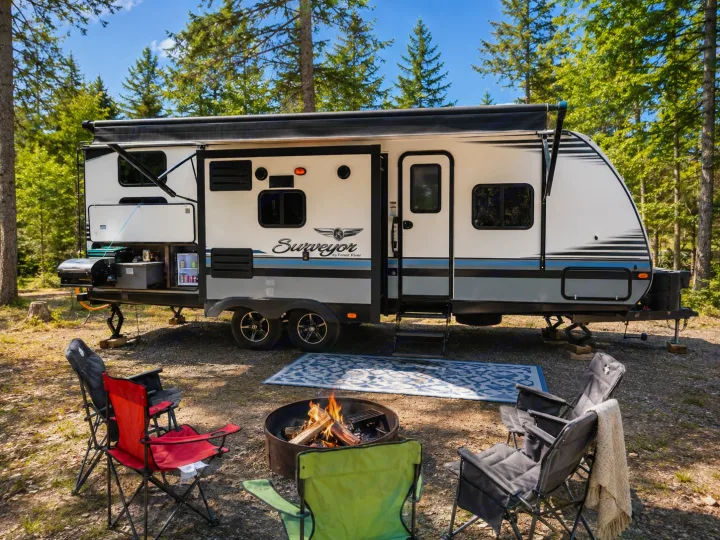 RV rental parked in a forest campsite, featuring an outdoor kitchen, warm campfire, and comfortable chairs for relaxation.. Forest River Surveyor 2018
