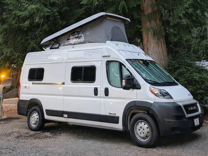 White Winnebago Solis pop-top campervan, an rv rental, parked on gravel in a lush, wooded area.. Winnebago Solis 2021