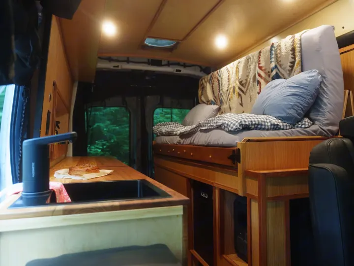 Inside an RV rental: a comfy bed, compact kitchenette with Dometic faucet & sink, and lush forest visible through the back.. Ford Transit 2025