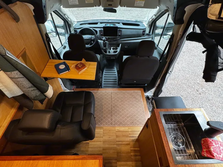 Experience van life in this RV rental: front cabin, a small kitchen with sink, and a cozy seating area with a table and pastry.. Ford Transit 2025