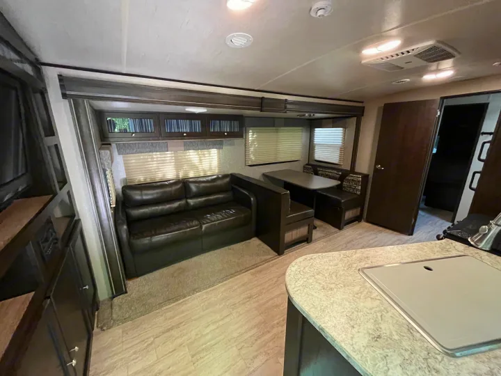 Inside an RV rental: a cozy living area with dark leather sofa, dining booth, light kitchen counter, and wood-look flooring.. Cruiser RV Shadow Cruiser 2018