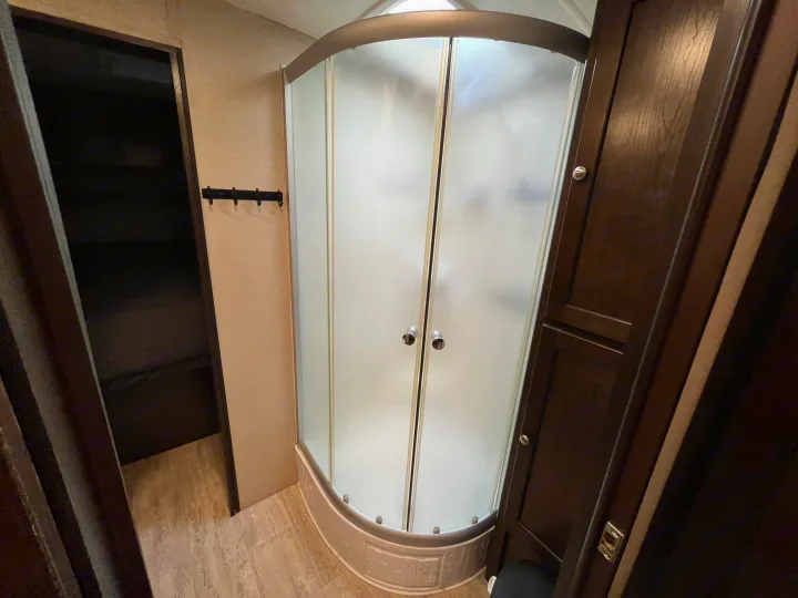 Inside an RV rental, this compact bathroom features a curved, frosted glass shower, dark wood cabinet, and wall hooks.. Cruiser RV Shadow Cruiser 2018
