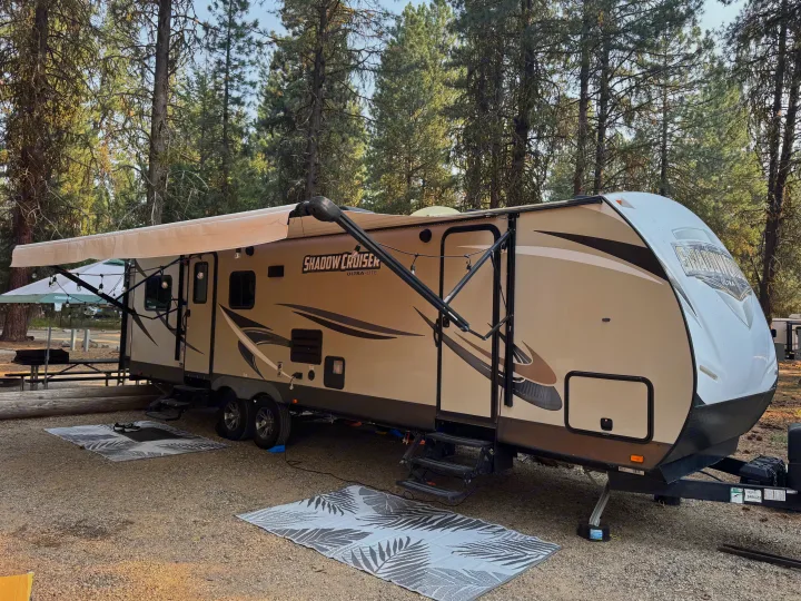 A 'Shadow Cruiser' RV rental is parked at a wooded campsite, featuring an extended awning with string lights and outdoor mats.. Cruiser RV Shadow Cruiser 2018