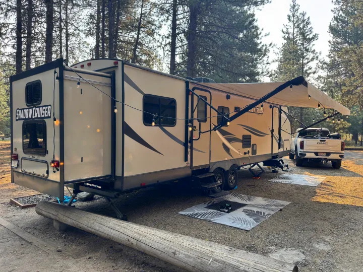 A Shadow Cruiser rv rental and truck parked in a serene forest campsite, complete with an extended awning and string lights.. Cruiser RV Shadow Cruiser 2018