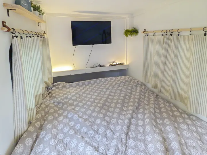 Compact RV rental bedroom featuring a bed with floral quilt, striped curtains, wall-mounted TV, and accent lighting.. MiniTrail Custom 2018