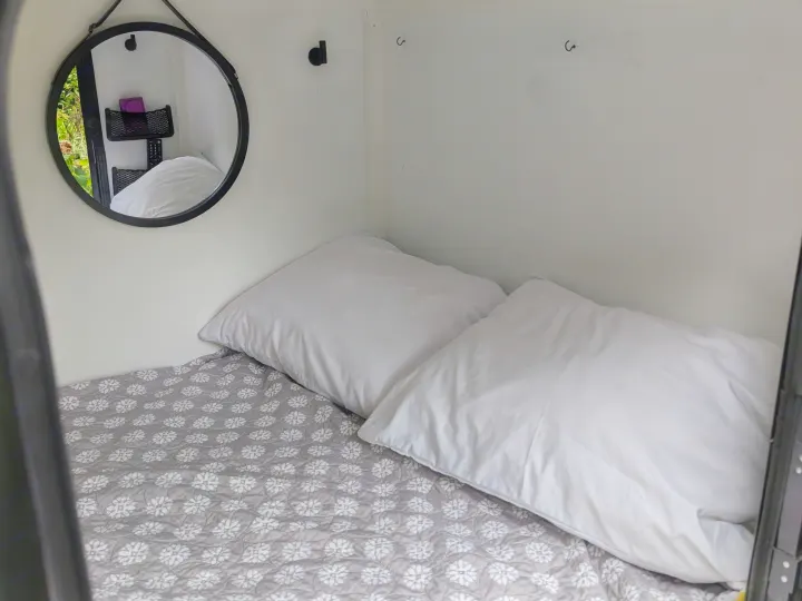 Cozy compact bed in an RV rental with white pillows, a grey floral blanket, and a mirror reflecting greenery.. MiniTrail Custom 2018