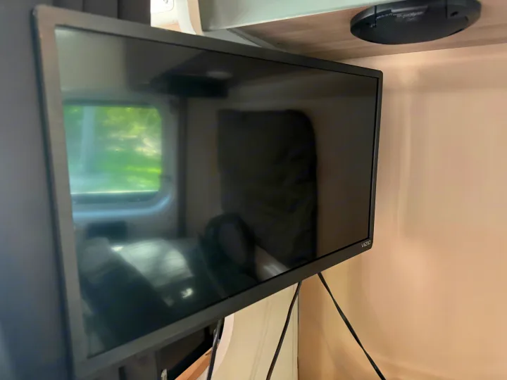 Vizio TV mounted in an RV rental, reflecting a window with green outdoor scenery. A speaker is above, with wires visible.. Dodge 3500shc 2021