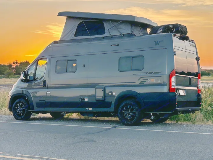 This dark grey RV rental camper van with a pop-top roof is parked roadside against a vibrant orange sunset.. Winnebago Solis 59PX 2027