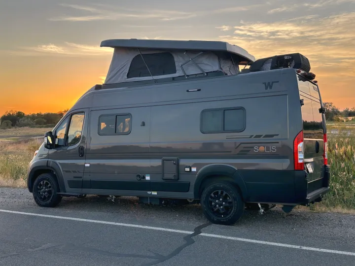 This gray Winnebago Solis pop-top camper van, ideal for rv rental, is seen at sunset by a grassy field.. Winnebago Solis 59PX 2027