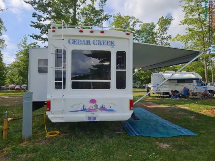 Sunny day at a campground featuring a white Cedar Creek **rv rental** travel trailer with an extended awning and scenic decal.. Cedar Creek Cedar Creek Fifth Wheel 2002