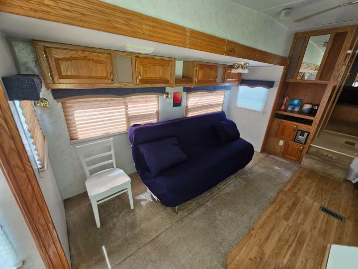 Inside an RV rental, showcasing a comfortable purple futon, white chair, and wood-toned cabinets above windows, defining a functional living area.. Cedar Creek Cedar Creek Fifth Wheel 2002