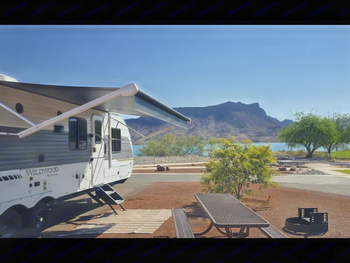 RV rental with an extended awning parked at a scenic lakeside campsite, featuring mountains, a picnic table, and a fire pit.. Forest River Wildwood X-Lite 2025