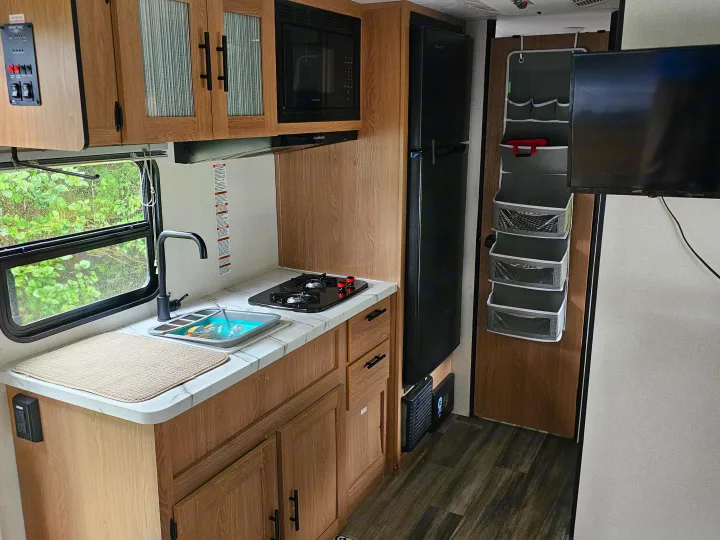 Compact rv rental kitchen interior showing wooden cabinets, a sink, stove, microwave, fridge, and a TV for your journey.. Avenger LT 2022