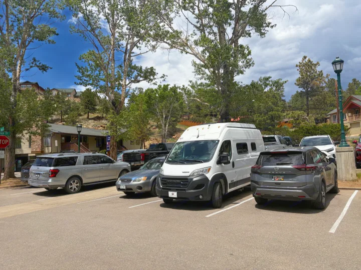 A white RV rental van parked in a lot surrounded by cars, trees, and hillside houses under a cloudy sky.. Winnebago Solis 2025